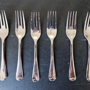 Puede incluir: Seis tenedores plateados dispuestos en fila. Cada tenedor tiene cuatro púas y un mango con un diseño en forma de corazón en la base. Los tenedores son de metal brillante y reflectante.