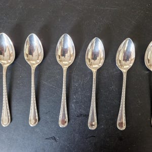 May include: A set of six silver-toned metal spoons arranged in a row. Each spoon features a polished, reflective oval bowl and a slender handle decorated with a delicate beaded border pattern that runs along the edges of the stem.