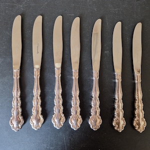 Könnte beinhalten: Acht silberne Essmesser mit verzierten Griffen. Die Messer haben eine polierte Oberfläche und sind in einer Reihe angeordnet.
