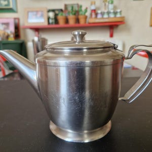 Puede incluir: Una tetera de acero inoxidable con tapa y asa curva. La tetera tiene un pico y se asienta sobre una base redonda. La tetera está sobre una mesa negra. El fondo está borroso.