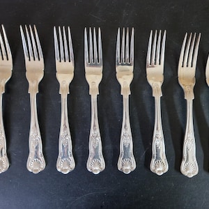 May include: Eight silver forks displayed in a line. Each fork features four tines and a decorative handle with an ornate floral and shell design. The forks are made of a shiny, reflective metal.