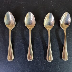 Könnte beinhalten: Vier goldfarbene Löffel mit ovalen Laffen und detaillierten Griffen sind auf einer dunklen Oberfläche angeordnet. Die Löffel sind gleichmäßig verteilt, reflektieren Licht und scheinen Teil eines Essgeschirrs zu sein.