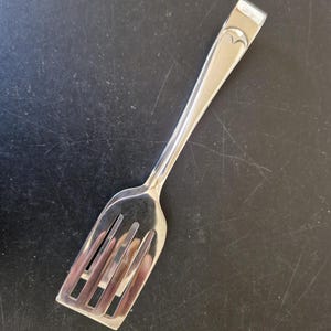 Puede incluir: Un tenedor de servir plateado con un diseño en forma de corazón en el mango. El tenedor tiene una cabeza rectangular con cuatro púas. El utensilio es de metal brillante y está sobre un fondo oscuro.