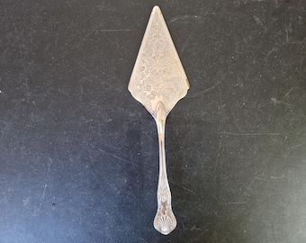 Könnte beinhalten: Ein Tortenheber aus silberfarbenem Metall mit einer dreieckigen Klinge, die ein detailliertes, geprägtes Blumen- und Rankenmuster aufweist. Der schlanke Griff läuft an der Basis in ein dekoratives, muschelartiges Motiv aus.