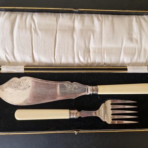 Puede incluir: Un juego de cuchillo y tenedor de mantequilla de plata con mangos de marfil, presentado en una caja forrada de terciopelo negro.
