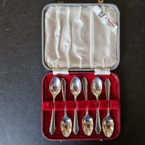 May include: A set of six silver-toned metal spoons arranged in a rectangular case with a red velvet interior and a white satin lid lining. The case features a label reading de Montfort Sheffield England and has a textured grey exterior finish.