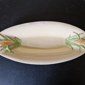 May include: A white ceramic serving dish with a raised design of corn on the cob on each end. The dish is oval shaped and has a cream coloured glaze.