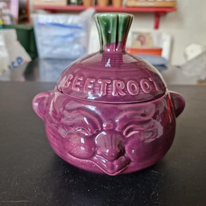May include: A purple ceramic beetroot-shaped container with a lid. The container has a smiling face design and the word "BEETROOT" is embossed on the lid. The stem is green. A unique kitchen decor item.