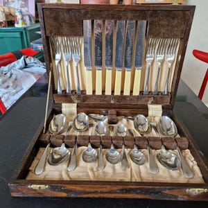Op de afbeelding: Een vintage houten bestekcassette, open om een complete set zilverwerk te onthullen. De set bevat vorken, messen met crèmekleurige handvatten en diverse lepels. De binnenkant is van fluweel.