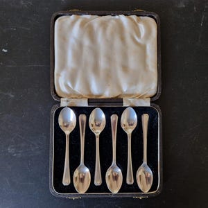 Puede incluir: Un juego de seis cucharas de plata presentadas en un estuche negro vintage con forro de satén crema. Las cucharas tienen cuencos redondeados y mangos largos y delgados. El estuche está abierto, mostrando las cucharas.
