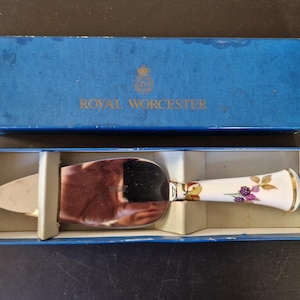 Może przedstawiać: Serwer do ciasta Royal Worcester w oryginalnym niebieskim pudełku. Serwer ma srebrne ostrze i białą rączkę ozdobioną złotymi wykończeniami, liśćmi i fioletowymi jagodami. Pudełko jest niebieskie ze słowami "Royal Worcester" wydrukowanymi na pokrywie.