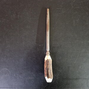 May include: A knife sharpener with a metal rod and a handle made of brown and white antler. The handle has a brass-colored ferrule. The sharpener is set against a dark background.