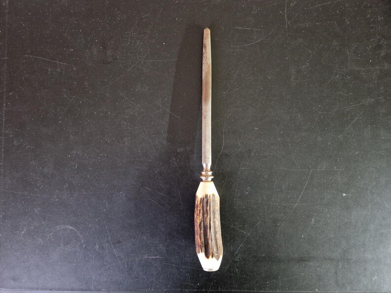 May include: A knife sharpener with a metal rod and a handle made of brown and white antler. The handle has a brass-colored ferrule. The sharpener is set against a dark background.