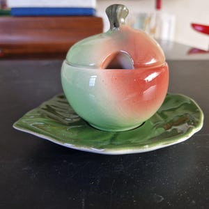 Pode incluir: Um açucareiro de cerâmica verde e rosa com uma tampa em forma de maçã. O açucareiro está sobre um prato verde em forma de folha.