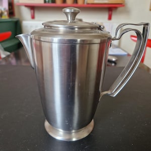 May include: A stainless steel coffee pot with a lid and a curved handle. The pot has a cylindrical body and a round base. The handle is attached to the side of the pot. The pot is sitting on a black surface.