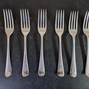 May include: Six silver forks with four tines each are arranged in a row on a dark surface. The forks have a simple, elegant design with a heart-shaped detail at the base of the handle. The tines are evenly spaced.