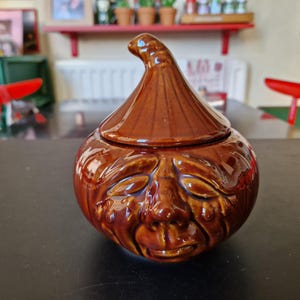 May include: A brown ceramic onion-shaped jar with a lid, featuring a face design. The jar has a glossy finish and is sitting on a dark surface. The lid has a pointed top.