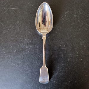 May include: A polished silver-toned metal spoon featuring an elongated oval bowl and a straight, slender handle that widens into a flat, rectangular end. The surface of the spoon is highly reflective, showing a smooth, metallic finish throughout.