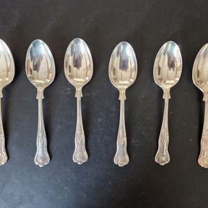 Op de afbeelding: Zes zilveren lepels gerangschikt op een donkere ondergrond. Elke lepel heeft een afgeronde kom en een decoratief handvat met een sierlijk ontwerp. De lepels reflecteren de omgeving.