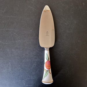Puede incluir: Un servidor de pastel de acero inoxidable con borde dentado. El mango es blanco con un diseño floral que presenta un melocotón y hojas verdes. El mango tiene un borde dorado en la base.