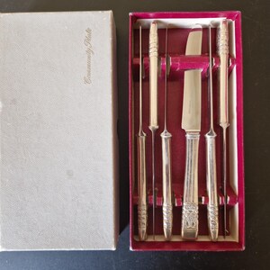 Könnte beinhalten: Ein silberfarbenes Besteckset in einer Box. Das Set enthält ein Messer und mehrere schlanke, spitze Werkzeuge, wahrscheinlich zum Servieren. Die Box ist hellgrau mit dem Aufdruck "Community Plate" auf der Vorderseite. Das Innere der Box ist mit rotem Samt ausgekleidet.