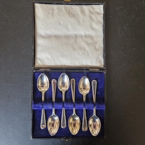 Puede incluir: Un juego de seis cucharaditas de plata en una caja forrada de terciopelo negro. Las cucharas están dispuestas en dos filas de tres, con los mangos hacia arriba. La caja está cerrada.
