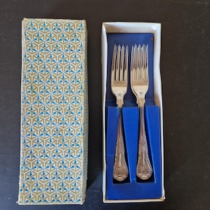 Peut inclure: Deux fourchettes argentées aux manches ornés sont présentées dans une boîte doublée de bleu. La boîte est ouverte, révélant les fourchettes nichées dans une boîte à motifs blancs et bleus. Les fourchettes sont en métal argenté.