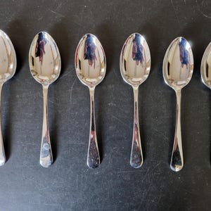 Puede incluir: Seis cucharas plateadas dispuestas en fila. Cada cuchara tiene un mango largo y delgado y un cuenco redondeado. Las cucharas están muy pulidas, reflejando el entorno. Las cucharas son probablemente para servir o comer.