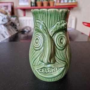 Może przedstawiać: Zielony ceramiczny kubek tiki z rzeźbionym wzorem twarzy. Twarz ma duże oczy, nos i usta z napisem "CELERY". Kubek ma teksturowany wygląd przypominający drewno.