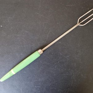 May include: A vintage fondue fork with a three-pronged stainless steel head and a light green handle. The handle has a white band near the end and a silver metal ferrule. The fork is approximately 25 cm long.