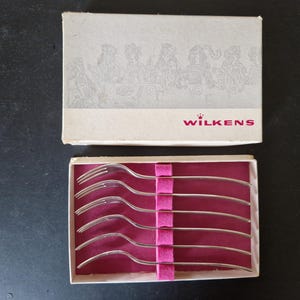 Puede incluir: Un juego de seis tenedores de plata en una caja blanca con interior rosa. La caja tiene el texto "WILKENS" impreso en la parte delantera.