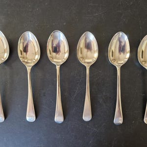 May include: A set of six polished silver-tone metal spoons arranged in a horizontal row. Each spoon features a smooth, rounded bowl and a plain, unadorned handle that tapers slightly toward the end, reflecting light on their metallic surfaces.