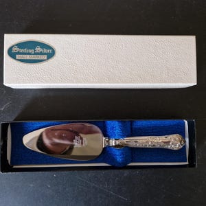 Peut inclure: Un couteau à gâteau en argent avec un manche orné dans une boîte bleue et noire. La boîte porte une étiquette blanche sur laquelle est inscrit "Sterling Silver Hall Marked".