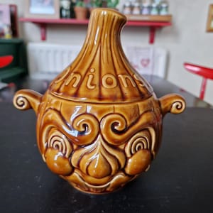 May include: Brown ceramic onion-shaped soup tureen with a lid. The tureen has a textured surface and the word "Onions" is written on the lid. It features handles and decorative swirls.