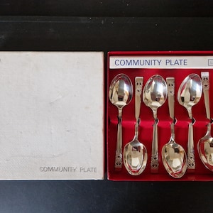 Pode incluir: Um conjunto de seis colheres prateadas numa caixa vermelha, com o texto "Community Plate" na caixa. As colheres têm um design decorativo nas pegas e estão dispostas ordenadamente na caixa. A caixa está aberta.