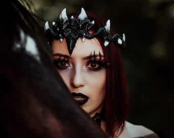 May include: A woman with red hair wears a black crown with white spikes. She has dark eye makeup and red contact lenses. She is looking at the camera, and a black horse's head is partially in the frame.