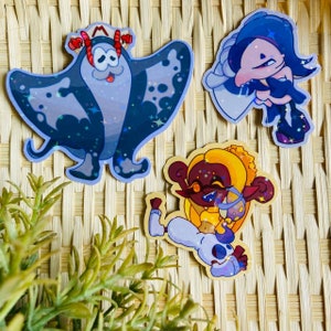 May include: Three colorful cartoon character stickers with glitter accents. The stickers feature a blue and purple manta ray, a yellow and purple character with a crown, and a blue and purple character with a black and white outfit.