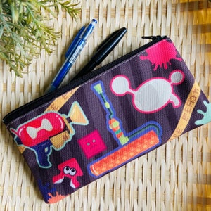 Puede incluir: Un estuche para lápices negro con cremallera con un estampado colorido de personajes y objetos de dibujos animados. El estampado incluye una pistola roja y rosa, una tubería azul y verde, una mancha blanca y un rodillo amarillo y naranja. El estuche está sobre una superficie de paja tejida.