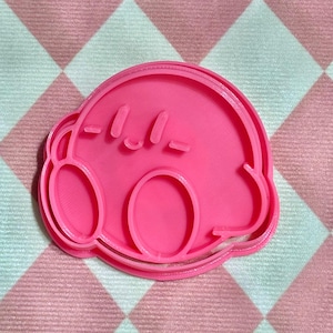 May include: Pink plastic cookie cutter shaped like the video game character Kirby. Kirby is a pink, round character with large eyes and a small mouth. The cookie cutter has a detailed outline of Kirby's body and face.
