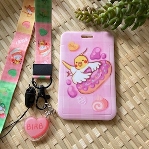 以下が含まれることがあります： ピンクのプラスチック製のIDカードホルダーで、黄色の鳥がピンクのドーナツを持っているという漫画のイラストが描かれています。カードホルダーとドーナツには「Birb」という文字が書かれています。カードホルダーの上部には、ネックストラップ用の穴があります。