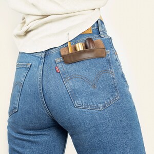 Könnte beinhalten: Eine Person trägt eine blaue Jeans mit einer braunen Ledertasche auf der rechten Gesäßtasche. Die Tasche enthält ein kleines Holzwerkzeug, ein goldenes Metallwerkzeug und ein kleines Holzwerkzeug.