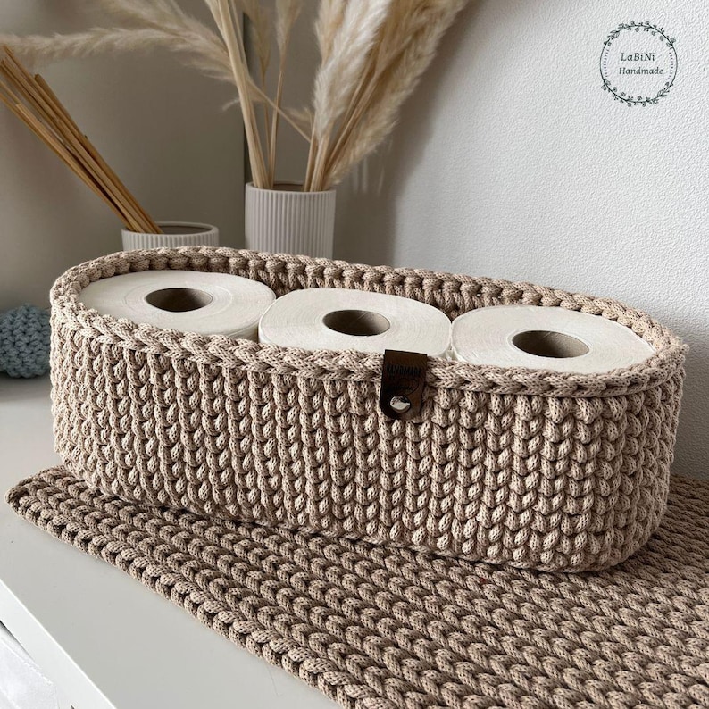 Puede incluir: Una cesta ovalada de ganchillo en color beige con una textura trenzada gruesa que contiene tres rollos de papel higi&eacute;nico blancos. La cesta presenta una peque&ntilde;a etiqueta de cuero marr&oacute;n con un remache met&aacute;lico en el borde frontal y descansa sobre una alfombrilla a juego.