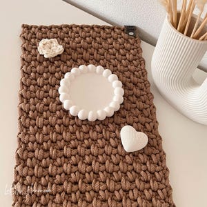 Könnte beinhalten: Eine rechteckige, braune Häkelmatte mit einem dicken, strukturierten Gewebe. Sie enthält eine kleine cremefarbene Häkelblume, ein weißes, rundes Tablett mit einem perlenartigen Rand und ein kleines weißes, herzförmiges Objekt mit Gitterstruktur.