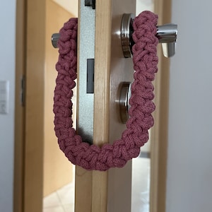 May include: A dusty rose colored, thick braided cord loop made of woven fabric. The U-shaped textile accessory is draped over a silver metal door handle attached to a light wood door, featuring a textured, interlocking knot pattern throughout.