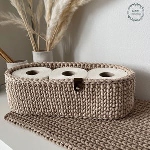Puede incluir: Una cesta de almacenamiento ovalada de ganchillo en color beige que contiene tres rollos de papel higi&eacute;nico blancos. La cesta presenta un patr&oacute;n de punto trenzado texturizado y una peque&ntilde;a leng&uuml;eta de cuero marr&oacute;n oscuro sujeta al borde frontal.