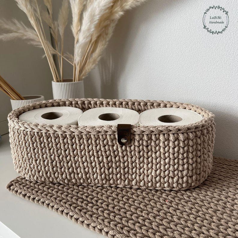 Puede incluir: Una cesta de almacenamiento ovalada de ganchillo en color beige que contiene tres rollos de papel higi&eacute;nico blancos. La cesta presenta un patr&oacute;n de punto trenzado texturizado y una peque&ntilde;a leng&uuml;eta de cuero marr&oacute;n oscuro sujeta al borde frontal.