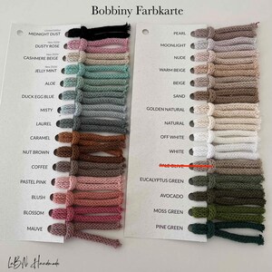 May include: A Bobbiny Farbkarte color chart featuring two white cards with rows of knotted textile cord samples in various shades including black, pink, beige, mint, blue, grey, brown, white, and green, with printed color names next to each sample.