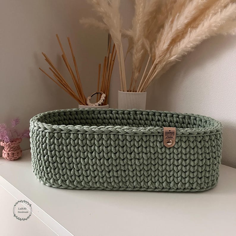 Puede incluir: Una cesta ovalada de ganchillo en color verde salvia hecha de hilo grueso. La cesta presenta un patr&oacute;n de punto trenzado visible y una peque&ntilde;a etiqueta rectangular de color canela en el lateral, sujeta con un remache met&aacute;lico y el texto Handmade.