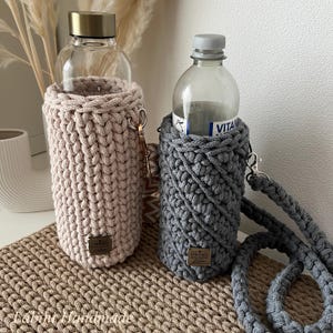 May include: Two cylindrical bottle holders made of thick crocheted cord. One holder is light beige with a vertical stitch pattern, and the other is dark gray with a diagonal stitch pattern. Both feature a small rectangular metal tag labeled Labini.