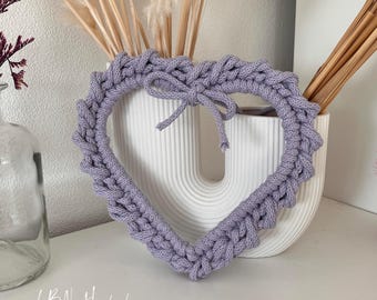 以下が含まれることがあります： 厚手のライトパープルのニット糸で作られたハート型の壁掛けで、上部に小さなリボンが付いています。この作品は、白いリブ状のU字型花瓶の前に配置されています。左下隅に「LaBiNi Handmade」というテキストが表示されています。